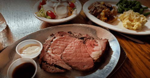 A plate with sliced prime rib, dipping sauces, and sides of salad, baked potato, and green beans on a wooden table.