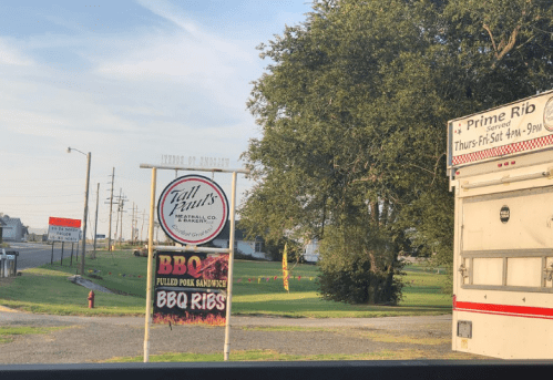 Sign for Tall Paul's Meat Market featuring BBQ ribs and pulled pork sandwiches, with a food truck nearby.