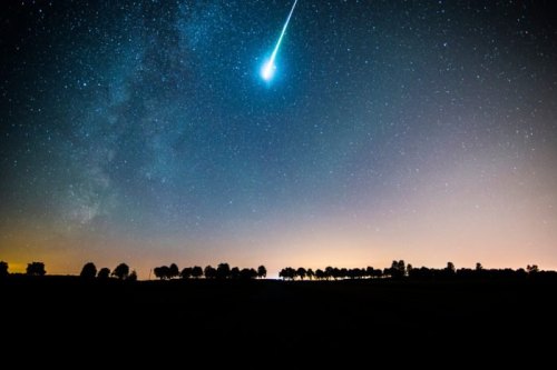 A bright meteor streaks across a starry night sky, with silhouettes of trees against a glowing horizon.