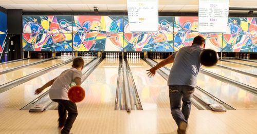 A child and an adult bowl together in a colorful bowling alley with vibrant murals and illuminated scoreboards.