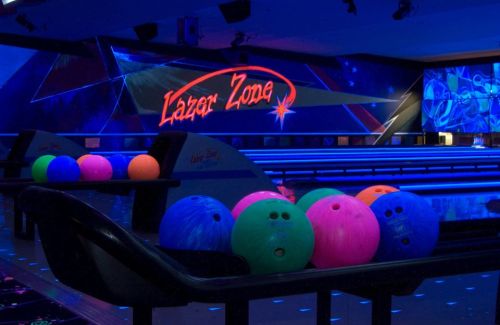 A vibrant bowling alley with neon lights, featuring colorful bowling balls on a rack and a "Lazer Zone" sign.