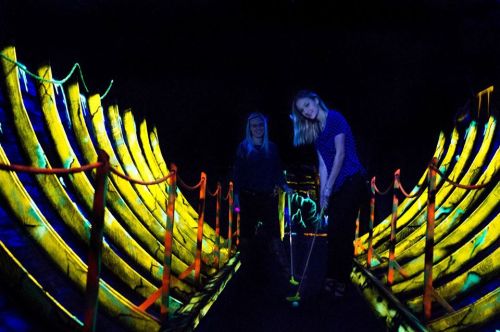 Two people play mini-golf in a brightly lit, neon-colored environment with glowing decorations.
