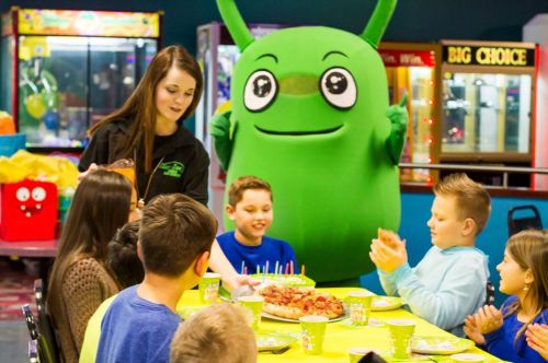 A green mascot stands behind a table of children celebrating a birthday with cake and snacks in a colorful party venue.