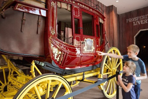 Two children examine a vintage red stagecoach with yellow wheels, pointing and discussing its details in a museum setting.
