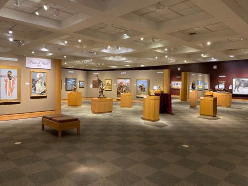 Art gallery interior featuring various paintings and sculptures displayed on wooden pedestals and walls.