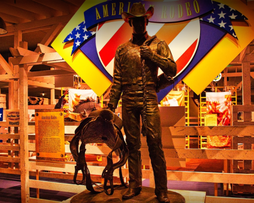 A bronze statue of a cowboy holding a saddle, set against a colorful rodeo-themed backdrop.