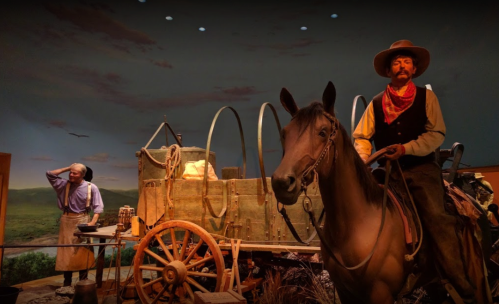 A cowboy on horseback next to a wagon, with another man in the background, set against a scenic backdrop.