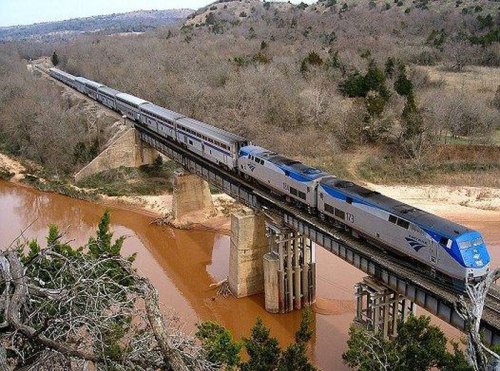 A train crosses a bridge over a river, surrounded by trees and hills in a rural landscape.