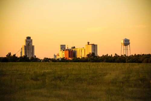 Sunset over a rural landscape featuring grain silos and a water tower, with a grassy field in the foreground.