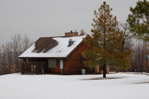 A wooden cabin surrounded by snow, with a pine tree nearby and a cloudy sky overhead.