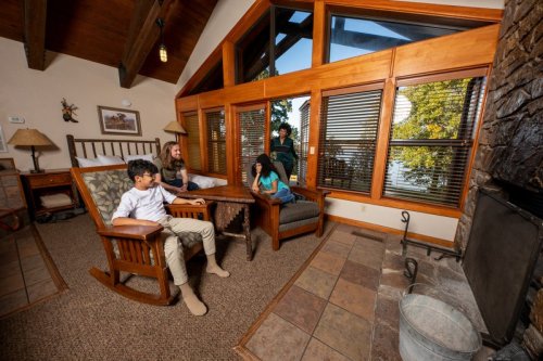 A cozy cabin interior with four people sitting and relaxing by a large window overlooking a lake.