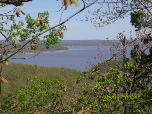 A serene view of a lake surrounded by lush greenery and distant hills under a clear blue sky.