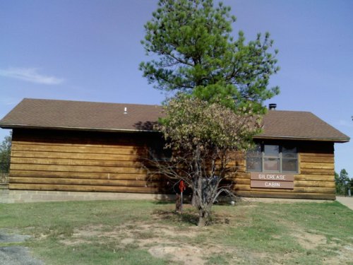 A wooden cabin with a sign reading "Gilcrease Cabin," surrounded by grass and a small tree.