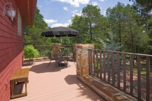 A cozy outdoor deck with a black umbrella over a table and chairs, surrounded by trees and blue skies.