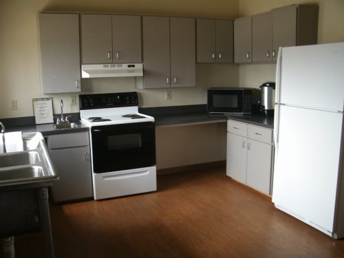 A simple kitchen with gray cabinets, a white stove, refrigerator, microwave, and a sink on a wooden floor.