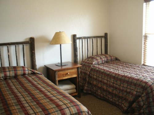 A cozy bedroom with two twin beds, a wooden nightstand, and a lamp, featuring plaid bedding and natural wood accents.