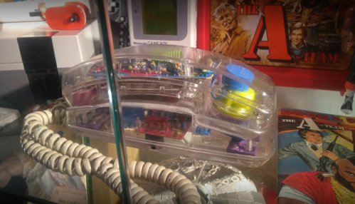 A clear, retro telephone with colorful buttons, displayed among vintage toys and memorabilia in a glass case.
