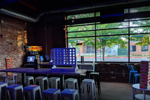 A cozy game room with a large window, featuring a Connect Four game, bar stools, and arcade machines.
