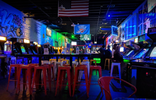A vibrant arcade bar with neon lights, arcade machines, and a lively atmosphere, featuring high-top seating and a bar area.