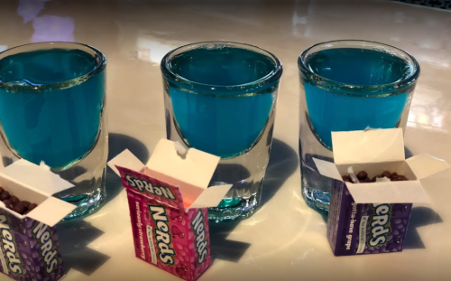 Three shot glasses filled with blue liquid, accompanied by opened boxes of Nerds candy in pink and purple.