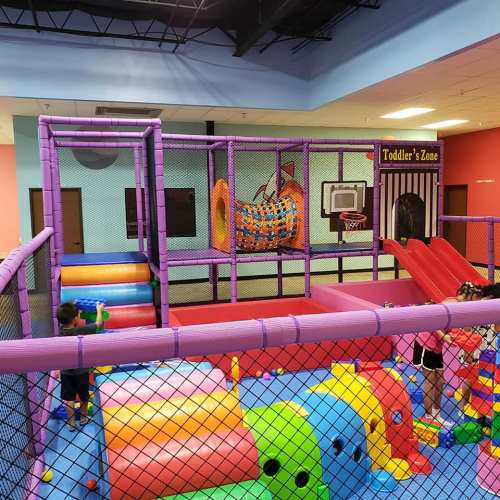 Colorful indoor play area with soft structures, slides, and a basketball hoop for toddlers.