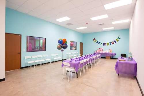 A brightly decorated birthday party room with a long table set with purple tablecloths and balloons.