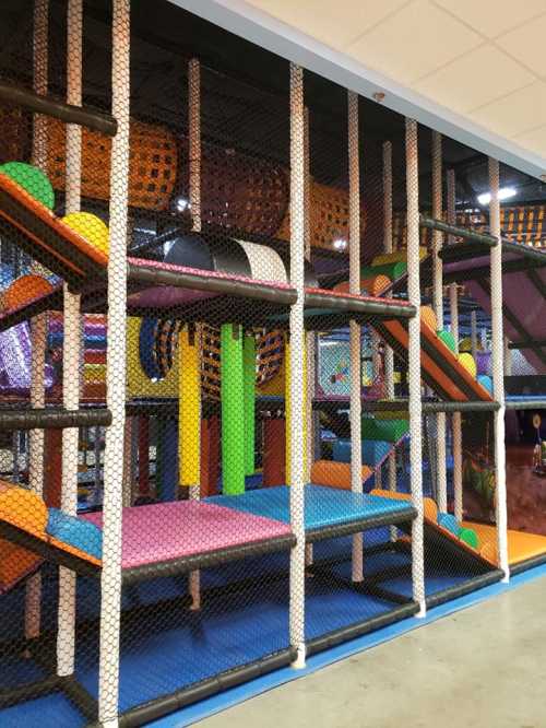 Colorful indoor playground with climbing structures, slides, and soft play areas enclosed by safety netting.