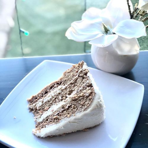 A slice of layered cake with white frosting on a square plate, next to a small vase with a white flower.
