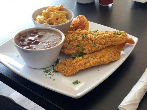 A plate with two fried fish fillets, a bowl of beans, and a side of macaroni and cheese, garnished with parsley.