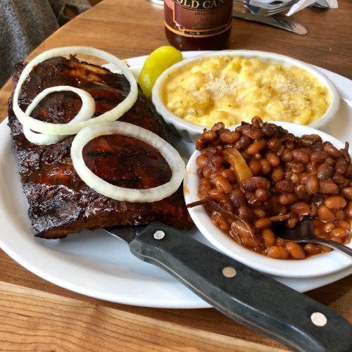 A plate featuring BBQ ribs with onions, macaroni and cheese, and baked beans, served with a pickle on the side.