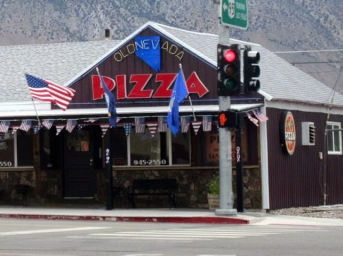 A rustic pizza restaurant with flags and a traffic light in front, set against a mountainous backdrop.