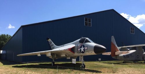 A military jet displayed outdoors in front of a blue hangar, with grass and a second aircraft in the background.