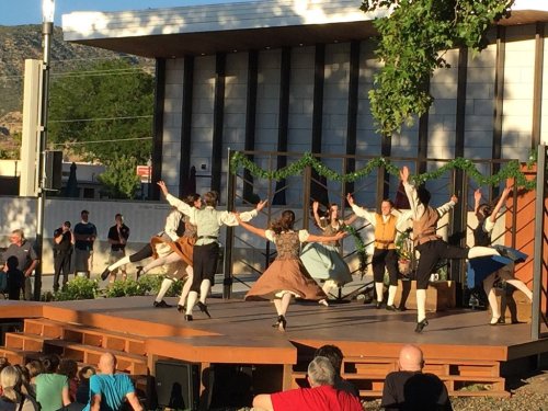 A lively outdoor dance performance with dancers in historical costumes, showcasing dynamic movements on a wooden stage.