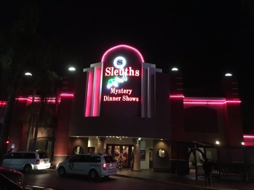 Exterior of Sleuths Mystery Dinner Shows at night, featuring bright neon signage and palm trees.