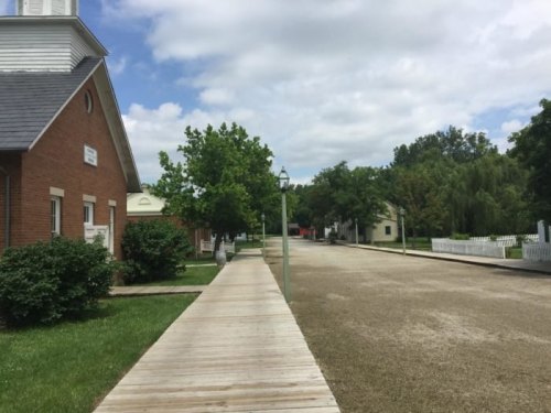 A quiet, tree-lined street with wooden walkways and historic buildings under a partly cloudy sky.