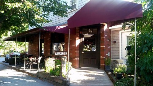 A cozy restaurant entrance with a maroon awning, welcoming sign, and outdoor seating surrounded by greenery.