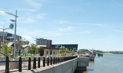 A scenic waterfront view featuring a walkway, trees, and buildings under a clear blue sky.