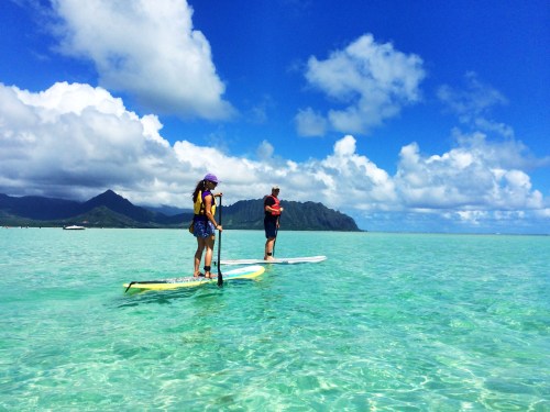 Two people paddleboarding on clear turquoise water with mountains and fluffy clouds in the background.