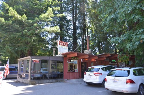 A cozy café surrounded by tall trees, featuring an outdoor seating area and several parked cars.