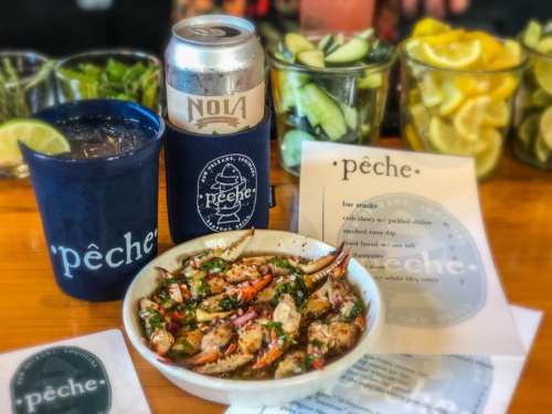 A bowl of seafood with crab claws, a can of beer, and two drinks on a bar counter, surrounded by fresh garnishes.