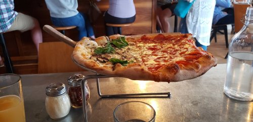 A large pizza on a metal peel, half cheese and half topped with mushrooms and basil, sits on a table in a restaurant.
