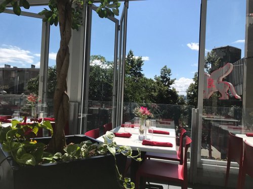 Bright restaurant interior with glass walls, tables set for dining, and greenery visible outside under a blue sky.