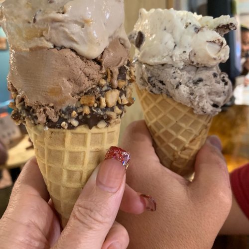 Two hands holding ice cream cones with different flavors, one topped with nuts and the other with cookie pieces.