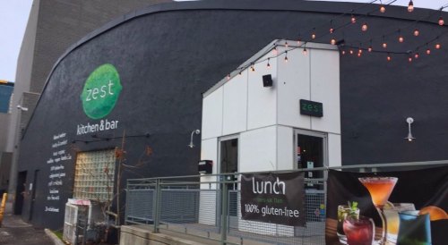 Exterior of Zest Kitchen & Bar, featuring a modern design, outdoor seating, and banners promoting gluten-free lunch options.