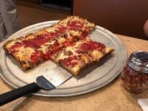A metal pizza tray with rectangular slices of pizza topped with cheese, pepperoni, and black olives, alongside a shaker of red pepper flakes.