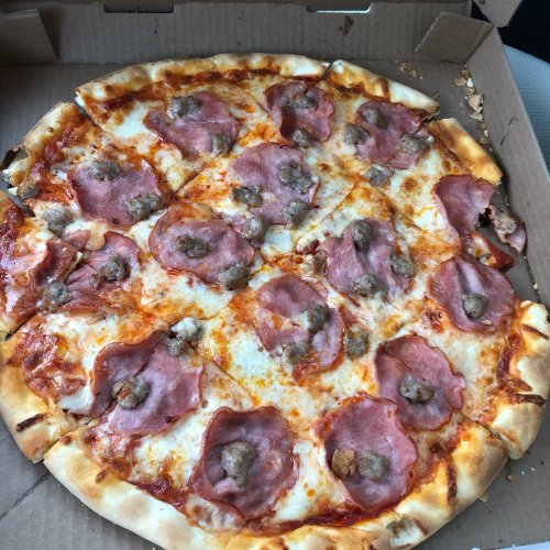A pizza with a golden crust topped with cheese, slices of ham, and small sausage pieces in a pizza box.