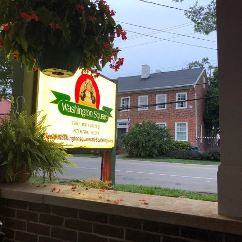 Sign for Washington Square Café and Catering, with a brick building in the background and hanging plants.