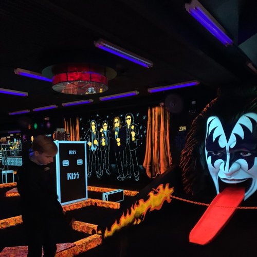 A vibrant indoor space featuring neon lights, a rock band mural, and a person in a costume with a striking face paint.