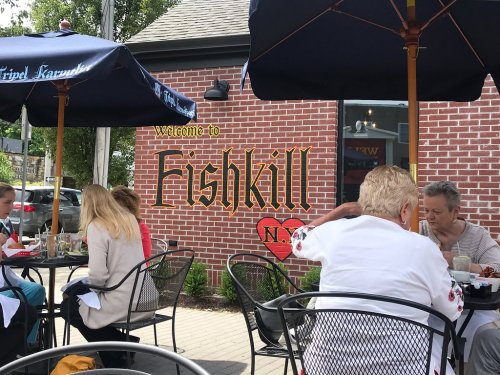 Outdoor dining scene with people at tables in front of a brick wall featuring a "Welcome to Fishkill, N.J." sign.