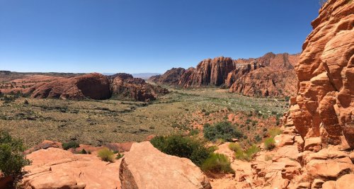 Panoramic view of red rock formations and a lush valley under a clear blue sky.